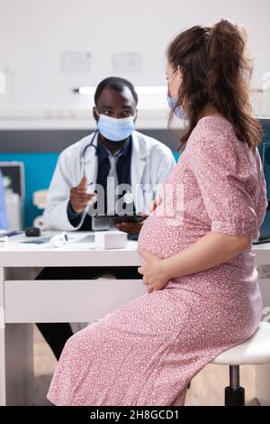 Médecin qui fait la consultation médicale avec la femme enceinte, portant des masques faciaux.Femme qui attend un enfant et qui reçoit des conseils médicaux du médecin au bureau pendant la pandémie Covid 19 Banque D'Images