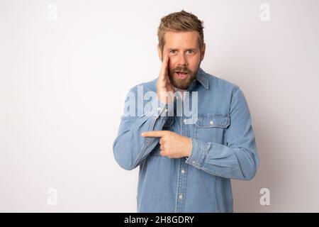 Jeune homme beau avec barbe portant une chemise en Jean sur fond blanc main sur la bouche en disant rumeur secrète, chuchotant malintentionnée conversation Banque D'Images
