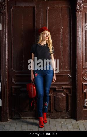 Image pleine longueur d'une femme à la mode dans une rue.Porter un Jean bleu skinny tendance, une chemise noire, des chaussures et un chapeau rouges. Banque D'Images