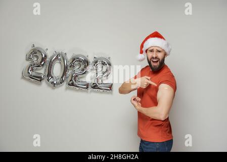 Un homme chauve avec une barbe dans un chapeau de Santa crie et montre biceps avec un plâtre après la vaccination contre le coronavirus (Covid-19) près de SIL poudré Banque D'Images