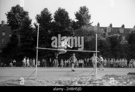 1964, historique, sports scolaires, heure d'été, Devon,L'Angleterre, le Royaume-Uni et l'extérieur à côté d'une fosse de sable traditionnelle, un jeune homme s'attaquant au saut en hauteur, un événement sur piste et terrain.Technique intéressante, ressemble à une forme du style Western Roll de saut où la jambe de plomb est levée en soulevant le corps qui tourne sur la barre avec la jambe de fuite repliée sous la plomb.Le Western Roll a finalement remplacé le saut en ciseaux original, dont il y avait beaucoup de variantes différentes, où le concurrent a généralement atterri sur leurs pieds. Banque D'Images