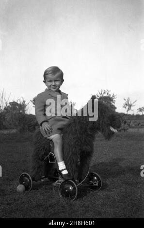 Années 1950, historique, à l'extérieur sur une certaine herbe, un petit garçon assis pour une photo sur un grand chien en peluche fourrure sur roues, Angleterre, Royaume-Uni.Les jouets à monter ou à pousser avec des roues étaient un jeu populaire pour les enfants de cette époque Banque D'Images