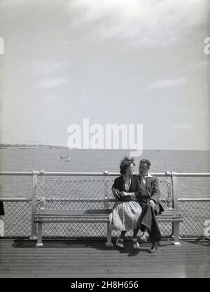 Années 1950, historique, un homme et une femme, de la fin des années vingt, dans les vêtements de l'époque, peut-être courting, assis près d'un banc en bois sur une jetée en bord de mer, Angleterre, Royaume-Uni. Banque D'Images