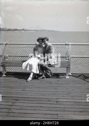 années 1950, historique, un homme et une femme assis étroitement ensemble sur un banc sur un quai en bois de bord de mer.De la fin des années vingt et le port des vêtements de l'époque, ils sont détendus, se tenant les mains, peut-être courting et/ou en vacances, Angleterre, Royaume-Uni. Banque D'Images