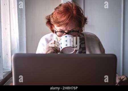 Femme âgée utilisant un ordinateur portable et buvant du café à la maison.Les personnes âgées surfent sur Internet, lisent des e-mails ou des actualités en ligne et naviguent sur les réseaux sociaux Banque D'Images