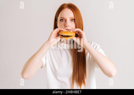 Portrait en gros plan d'une charmante jeune femme en train de savourer un délicieux hamburger appétissant en regardant l'appareil photo sur fond blanc et isolé en studio. Banque D'Images