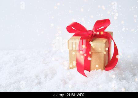 Boîte cadeau artisanale ornée d'un noeud rouge dans la neige.Fond blanc et lumières de fête. Banque D'Images