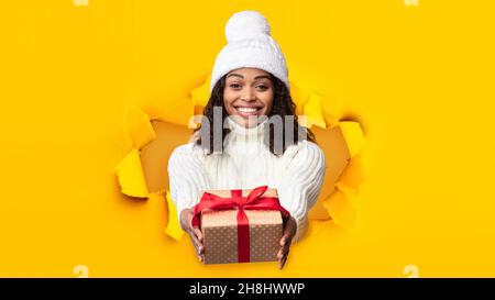 Joyeux millénaire jolie femme noire en chapeau blanc et chandail tient la boîte cadeau dans le trou dans le papier jaune Banque D'Images