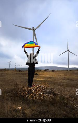 Femme debout sur une petite colline, son dos méconnaissable élevant un drapeau arc-en-ciel regardant vers l'horizon bleu et nuageux, dans un parc de vent de tur Banque D'Images