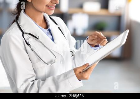 Pantacourt souriant jeune jolie femme indienne médecin en manteau blanc examine les antécédents médicaux et consulte sur comprimé Banque D'Images