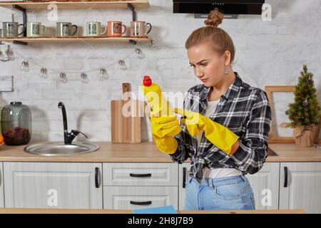 Jeune femme professionnelle portant des gants de protection en caoutchouc jaune tenant des nettoyants pour bouteille dans la cuisine. Banque D'Images