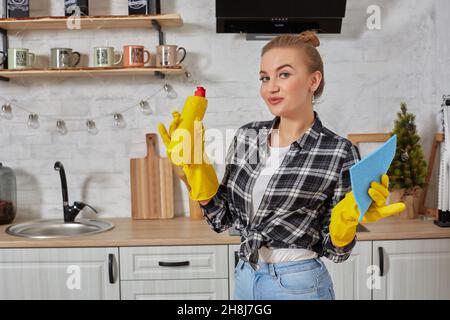 Jeune femme professionnelle portant des gants de protection en caoutchouc jaune tenant des nettoyants pour bouteille dans la cuisine. Banque D'Images