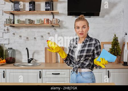 Jeune femme professionnelle portant des gants de protection en caoutchouc jaune tenant des nettoyants pour bouteille dans la cuisine. Banque D'Images