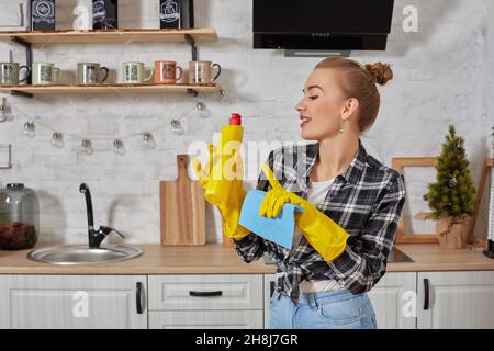 Jeune femme professionnelle portant des gants de protection en caoutchouc jaune tenant des nettoyants pour bouteille dans la cuisine. Banque D'Images