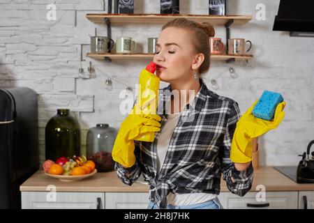 Jeune femme professionnelle portant des gants de protection en caoutchouc jaune tenant des nettoyants pour bouteille dans la cuisine. Banque D'Images