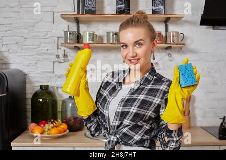 Jeune femme professionnelle portant des gants de protection en caoutchouc jaune tenant des nettoyants pour bouteille dans la cuisine. Banque D'Images