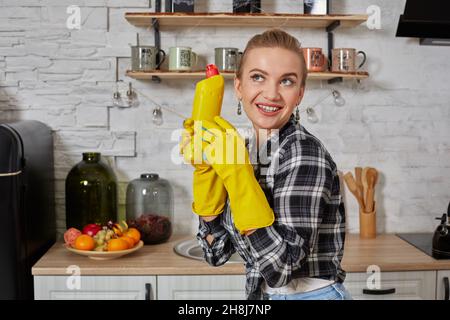 Jeune femme professionnelle portant des gants de protection en caoutchouc jaune tenant des nettoyants pour bouteille dans la cuisine. Banque D'Images