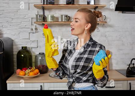 Jeune femme professionnelle portant des gants de protection en caoutchouc jaune tenant des nettoyants pour bouteille dans la cuisine. Banque D'Images