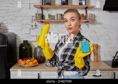 Jeune femme professionnelle portant des gants de protection en caoutchouc jaune tenant des nettoyants pour bouteille dans la cuisine. Banque D'Images