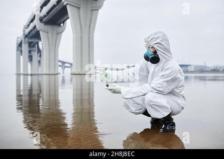 Vue latérale portrait en longueur d'un scientifique portant une combinaison de matières dangereuses pour la collecte d'échantillons d'eau, mains tenant le tube à essai, espace de copie Banque D'Images