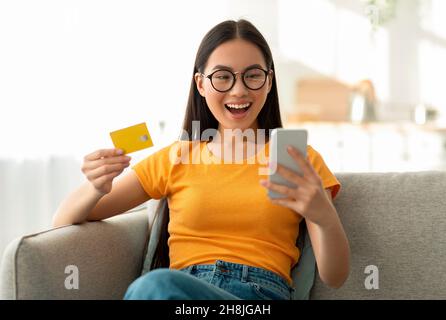 Une jeune femme asiatique surjoyeuse en lunettes achetant sur le Web à la maison, assis sur un canapé, en utilisant un smartphone et une carte de crédit Banque D'Images