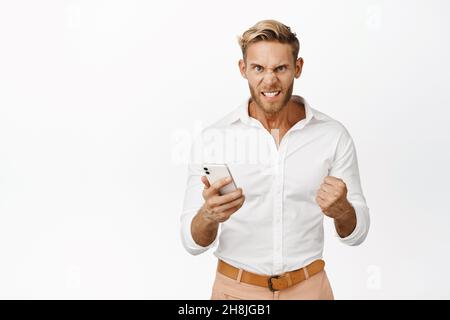 Homme blond en colère tenant un téléphone portable, un poing furieux et contrarié, debout sur fond blanc Banque D'Images