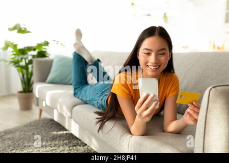 Sentiments agréables d'achat.Bonne femme asiatique regardant un smartphone et utilisant une carte de crédit, allongé sur un canapé et souriant Banque D'Images