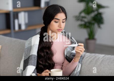 Bouleverser la jeune femme hindoue avec une tasse de boisson chaude dans le plaid, souffrant de froid, assis sur le canapé dans la salle de séjour Banque D'Images