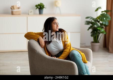 Une dame du Moyen-Orient rêveuse assise dans un fauteuil confortable et donnant sur l'extérieur, appréciant une matinée tranquille, ayant un week-end paisible Banque D'Images