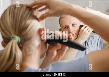 L'homme qui se rasante murmure les cheveux lui-même, coupe-cheveux à la maison devant le miroir dans le bain.Homme avec une coupe longue à l'aide de la tondeuse pour machine à rasage Banque D'Images