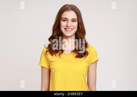 Portrait d'une adolescente gaie en jaune T-shirt décontracté sincèrement souriant à l'appareil photo, humeur positive, être heureux de rencontrer quelqu'un.Prise de vue en studio isolée sur fond gris. Banque D'Images