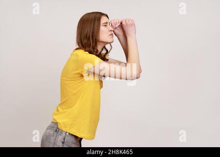 Vue latérale d'une jeune fille aux cheveux bruns en jaune T-shirt décontracté regardant à travers les jumelles gestuelle, zoom de la vision, exploration de la distance.Prise de vue en studio isolée sur fond gris. Banque D'Images