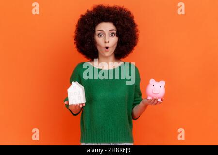 Femme surprise avec la coiffure d'Afro portant vert décontracté style chandail tenant la banque de piggy et la maison de papier dans les mains, l'achat de propriété.Studio d'intérieur isolé sur fond orange. Banque D'Images