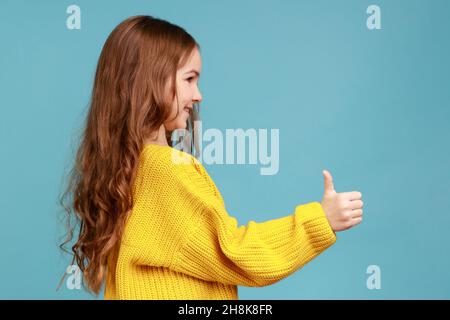 Vue latérale portrait de petite fille charmante mignonne debout avec le pouce vers le haut, montrant le geste approuvé, portant jaune style décontracté pull.Studio d'intérieur isolé sur fond bleu. Banque D'Images