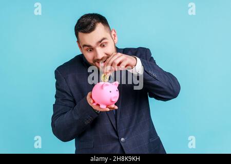 Homme enthousiaste avec une barbe portant un costume de style officiel, investissant dans des bitcoins, mettant la pièce de crypto dorée dans la banque de piggy, heureux avec des économies d'argent.Studio d'intérieur isolé sur fond bleu. Banque D'Images