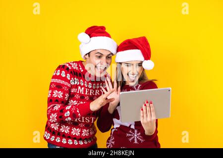 jeune couple latin ayant des appels vidéo de noël à l'aide d'une tablette numérique ou d'un pavé tactile sur un fond jaune en amérique latine Banque D'Images