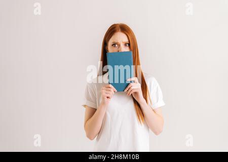 Portrait en studio de jeune étudiante effrayée cache le visage derrière l'ordinateur portable regardant l'appareil photo, debout sur un arrière-plan blanc isolé. Banque D'Images