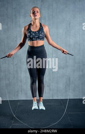 Gaie athlétique fille dans sportswear femme entraînement avec une corde à sauter dans une salle de gym sur un mur gris. Banque D'Images