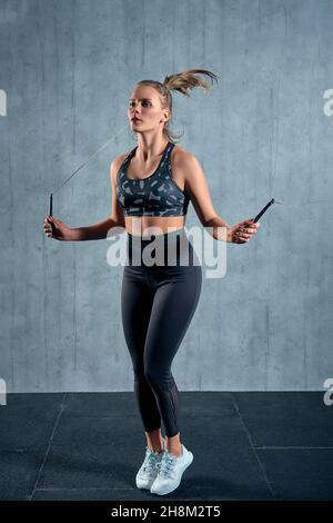 Gaie athlétique fille dans sportswear femme entraînement avec une corde à sauter dans une salle de gym sur un mur gris. Banque D'Images