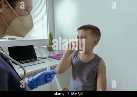 Enfant ayant un test PCR Saline Gargle effectué au cabinet du médecin.Femme médecin aidant un garçon de 7 ans avec le test Covid-19 PCR Gargle à la clinique médicale Banque D'Images