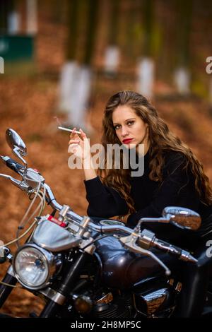 Une belle femme aux cheveux longs fumant sur une moto hache en paysage d'automne sur une route forestière Banque D'Images