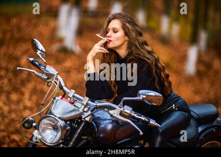 Une belle femme aux cheveux longs fumant sur une moto hache en paysage d'automne sur une route forestière Banque D'Images