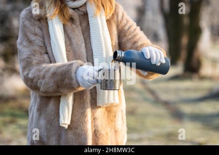 La femme verse une boisson chaude des thermos dans une tasse en métal.Rafraîchissement dans la nature hivernale.Femme porte un manteau de fourrure, une écharpe tricotée et des gants. Banque D'Images