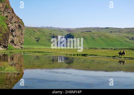 Les masses d'eau s'effondrent verticalement dans les profondeurs, se reflètent dans l'eau, paysage vert, deux cavaliers sur les chevaux islandais, Skogafoss, Islande Banque D'Images