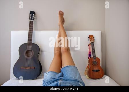 Femme avec instruments au lit. Fille avec sa guitare et son ukulele sur le lit dans sa chambre à la maison. Guitare espagnole. Banque D'Images