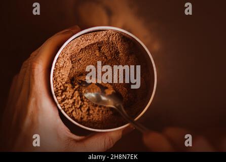 Une femme tient dans ses mains un pot rempli de poudre de cacao naturel parfumé, qu'elle prend avec une cuillère à café.Le processus de fabrication d'un délicieux inv Banque D'Images