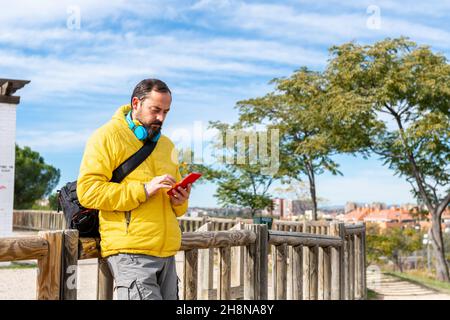 Un petit boxeur sérieux avec une barbe vêtue d'un manteau jaune, un sac à bandoulière à l'arrière et un casque sans fil bleu discutant avec son téléphone portable Banque D'Images