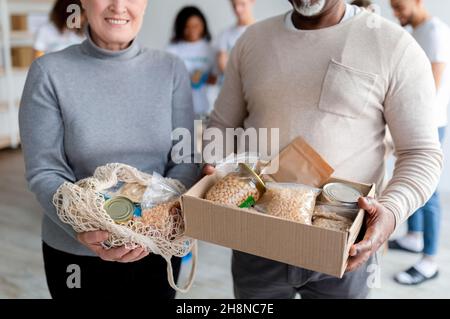 Les conjoints âgés de race mixte montrant des dons alimentaires dans une boîte et un sac à ficelle, les volontaires emballant des boîtes pour les pauvres Banque D'Images