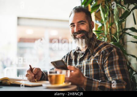 L'homme adulte est assis dans un café et fait son horaire quotidien.Il utilise un smartphone et un bloc-notes. Banque D'Images