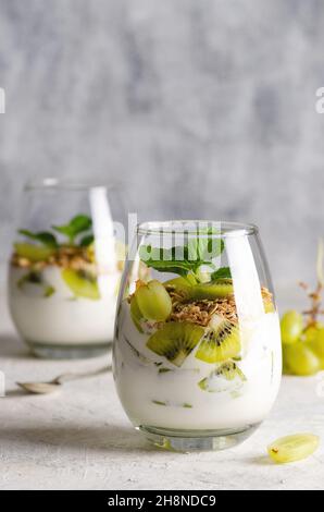 Deux verres de yaourt nature avec des morceaux de kiwis frais, des raisins blancs, des céréales et des feuilles de menthe sur fond gris. Banque D'Images